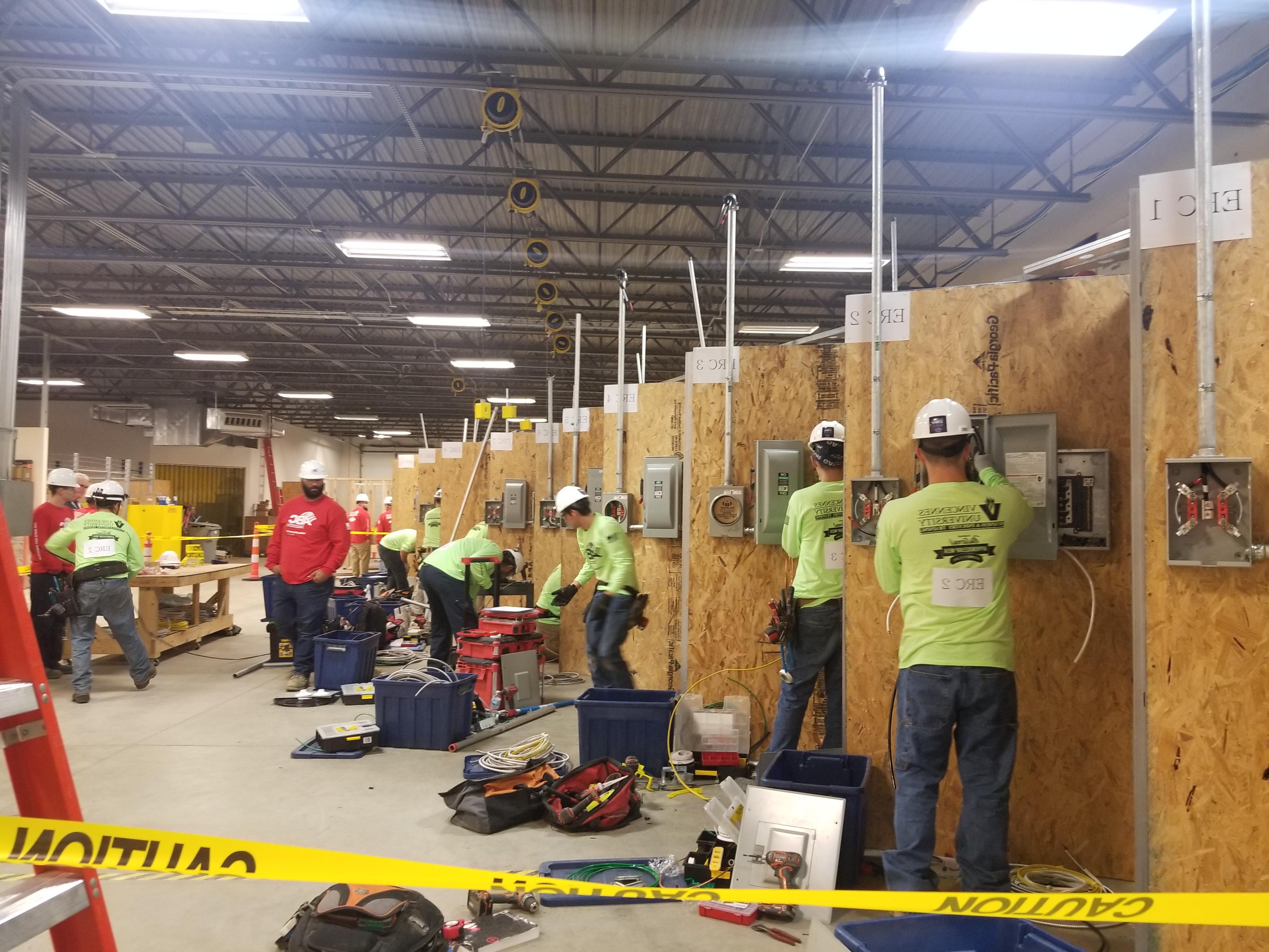 People in hard hats work at individual electrical panels with equipment around them while Instructors look on.