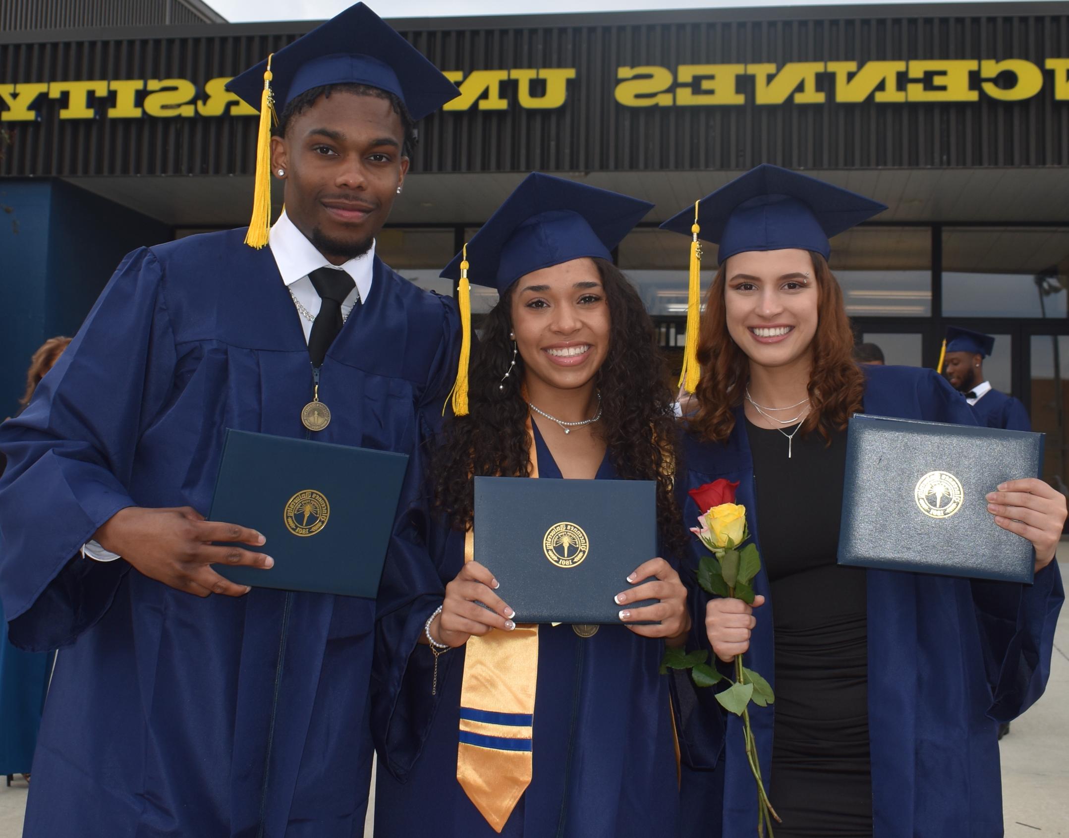 两名女学生和一名男学生戴着毕业帽，穿着毕业服，拿着学位，身后是文森斯大学的标志.