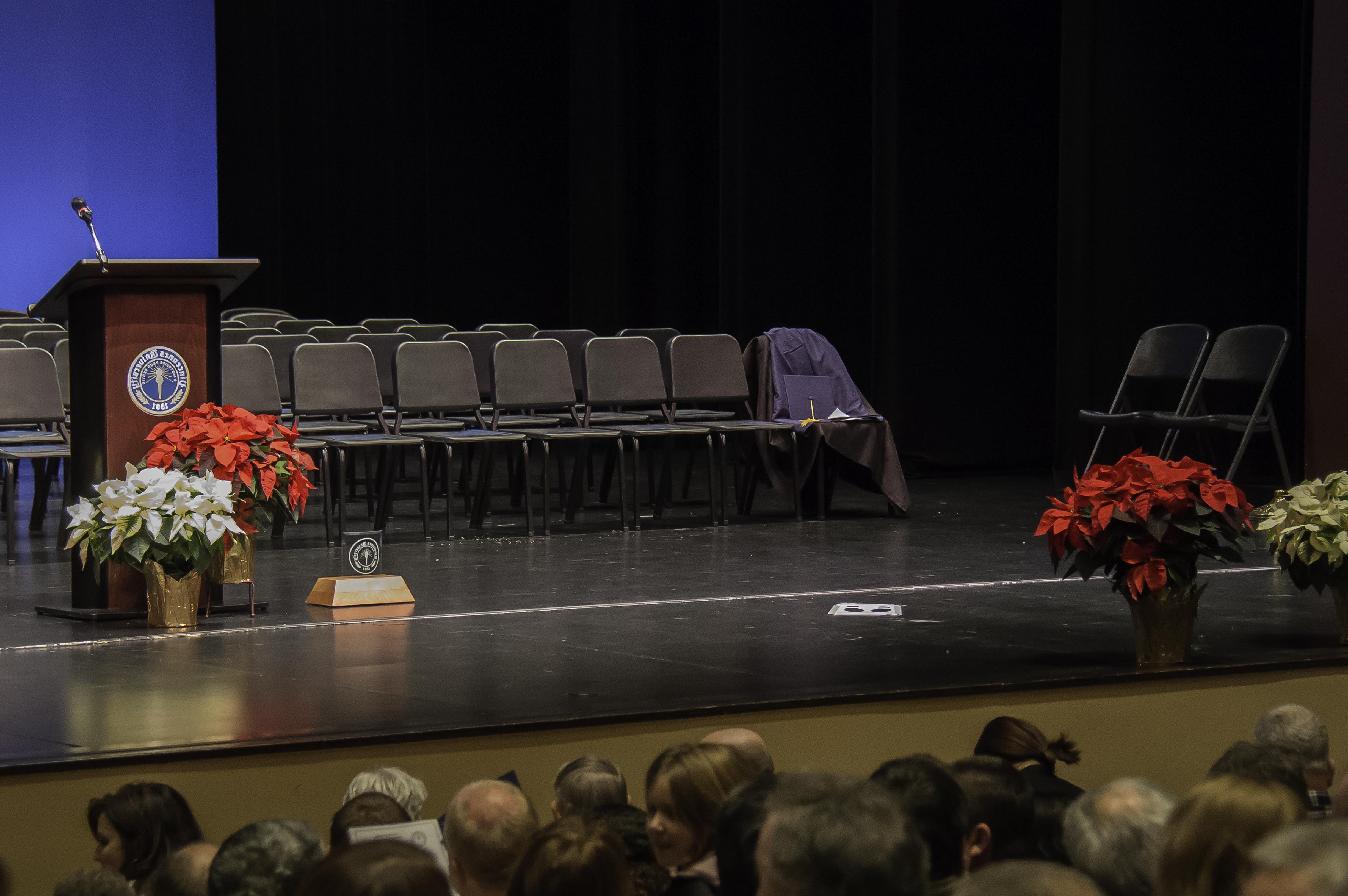 椅子，一品红，红骷髅表演艺术中心舞台上的讲台. 其中一把椅子上放着一顶帽子和一件长袍. 男嘉宾和女嘉宾坐在舞台前的观众席上. 